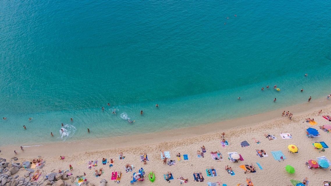 Una vista aerea di una spiaggia sabbiosa e affollata dove le persone prendono il sole su teli colorati e nuotano nel limpido mare turchese.