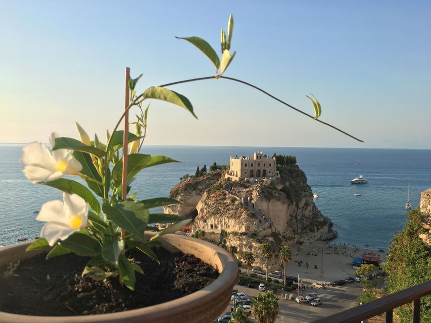 Un monastero si erge su un'isola rocciosa, affacciato su una spiaggia e il mare, visto da un balcone con una pianta in vaso.