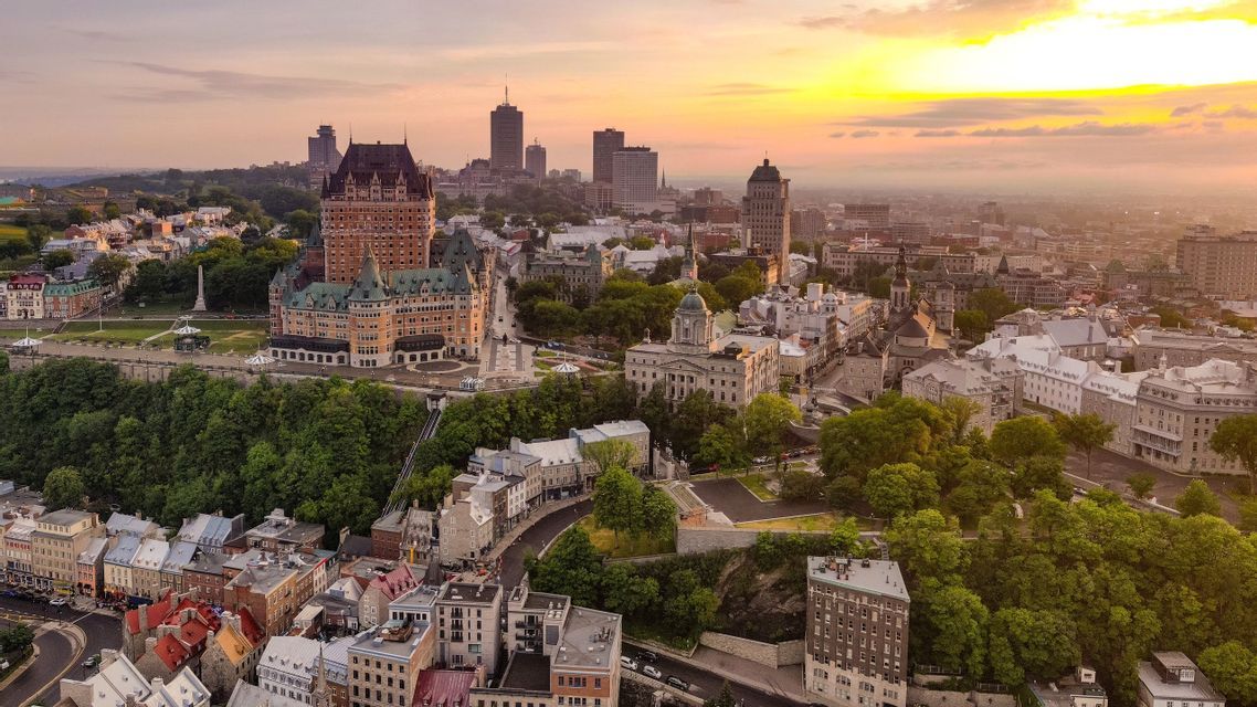 Vue aérienne d'une ville avec un grand hôtel aux allures de château et des bâtiments historiques nichés parmi les arbres, sous un coucher de soleil doré.
