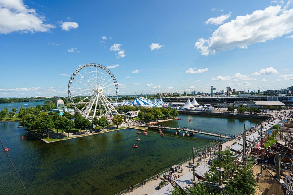 Une grande roue blanche et des tentes de cirque bordent un parc au bord de l'eau où des gens se promènent et font du bateau sous un ciel bleu.