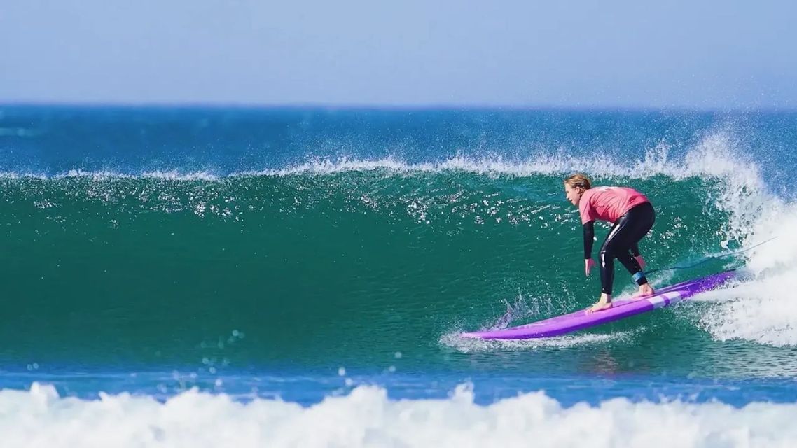 Une personne en chemise rose et combinaison de plongée surfe sur un longboard violet, chevauchant une grande vague turquoise de l'océan sous un ciel dégagé.