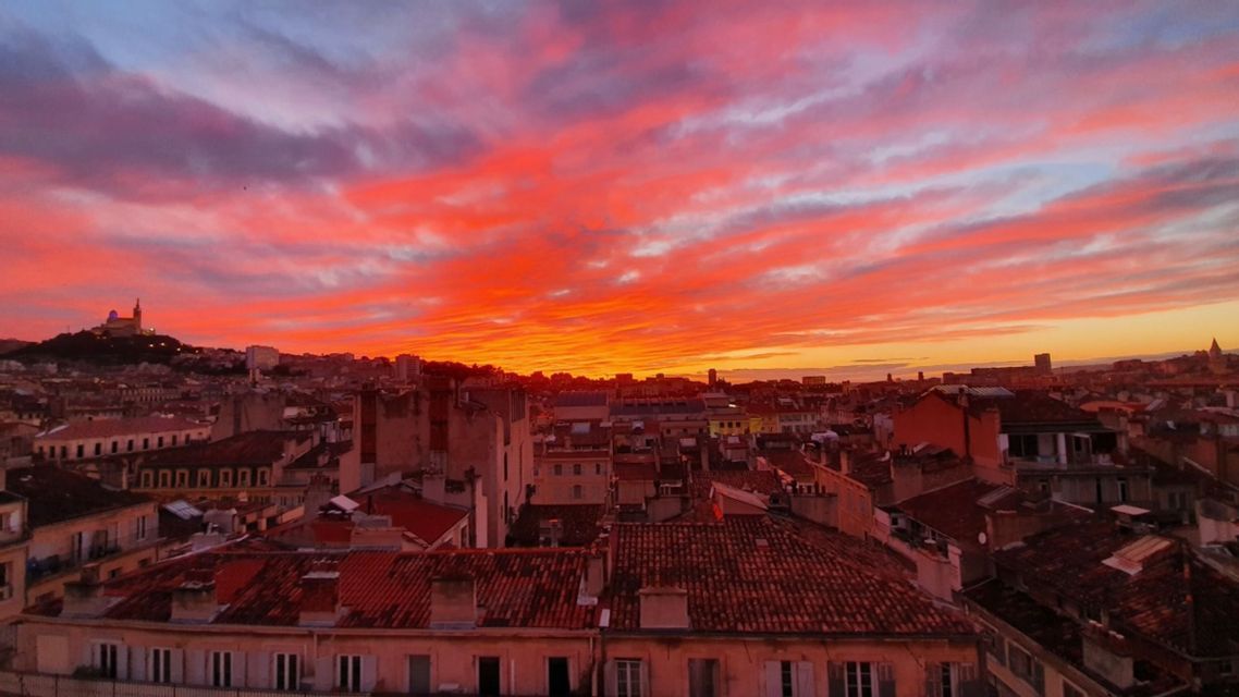 Un paysage urbain tentaculaire avec des toits en terre cuite illuminés par un coucher de soleil vibrant, orange et rouge.