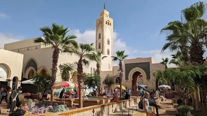 Une place de marché en plein air animée avec un haut minaret, des palmiers et des passants déambulant parmi les étals sous un ciel bleu et ensoleillé.