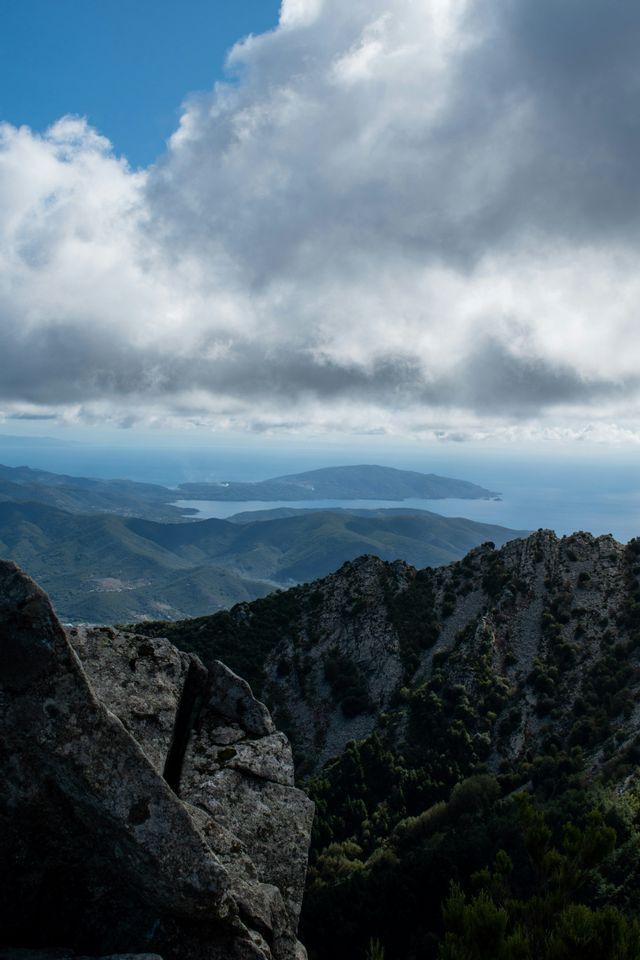 Vista da una cima montuosa rocciosa che si affaccia su verdi colline, un'isola e il mare sotto un cielo azzurro nuvoloso.