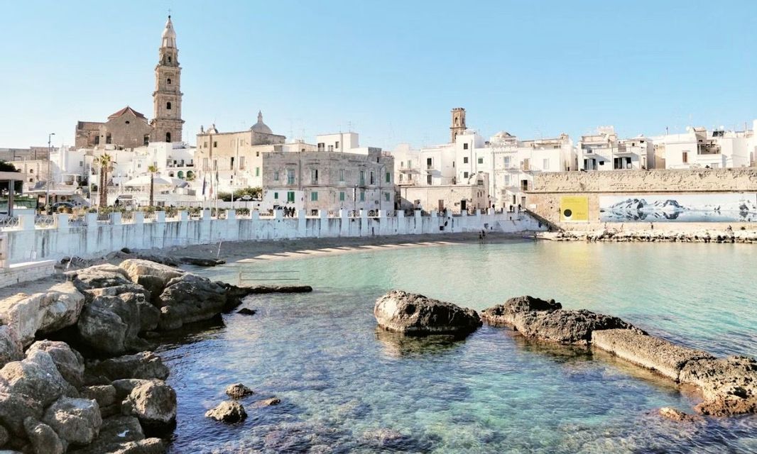 Vista soleggiata di una storica città costiera con edifici bianchi e un campanile, da una limpida baia turchese.