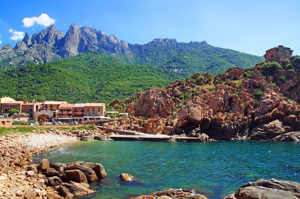 A coastal village sits on a rocky bay with turquoise water, with large green mountains rising in the background under a blue sky.