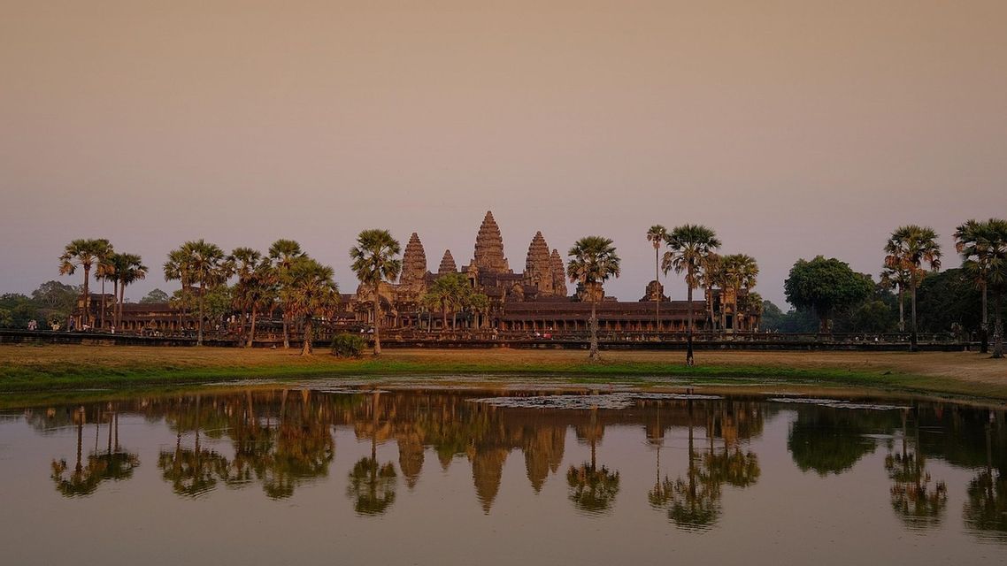 Un ancien complexe de temples avec des tours et des palmiers se reflète dans l'eau calme d'un lac au coucher du soleil.