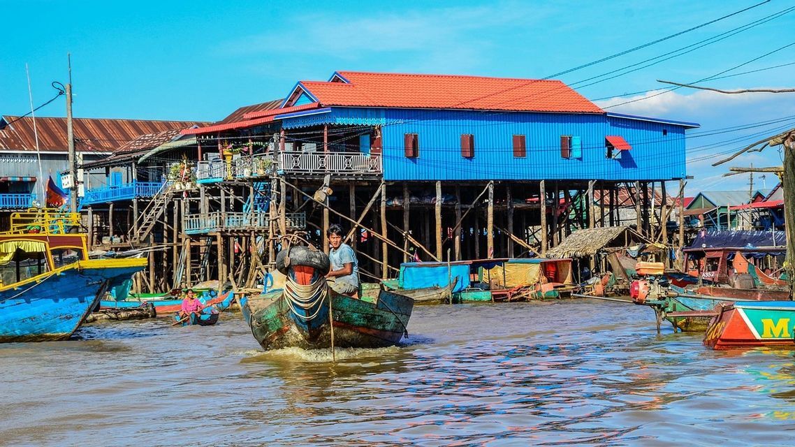 Un homme dans un bateau en bois navigue sur une rivière le long de maisons colorées sur pilotis dans un village flottant sous un ciel bleu clair.