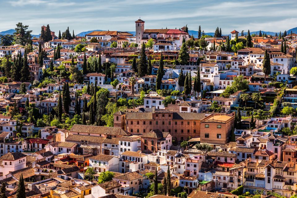 Una vista panorámica de una ciudad construida en una ladera, con muchos edificios blancos, tejados de terracota y altos cipreses.