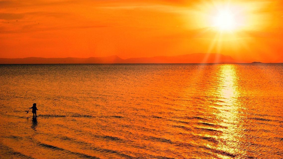 Une silhouette d'une personne debout dans la mer peu profonde au coucher de soleil orange éclatant, le soleil se reflétant sur l'eau.