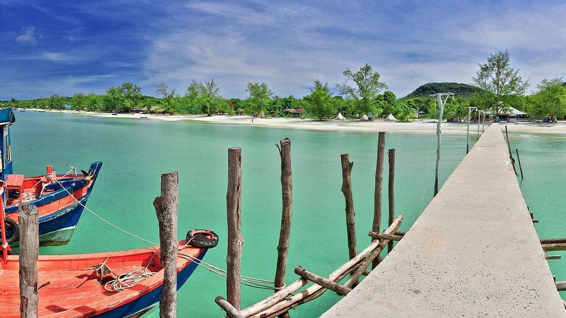 Une jetée en béton s'étire sur des eaux turquoises vers une plage de sable bordée d'arbres luxuriants, où un bateau rouge et bleu est amarré.