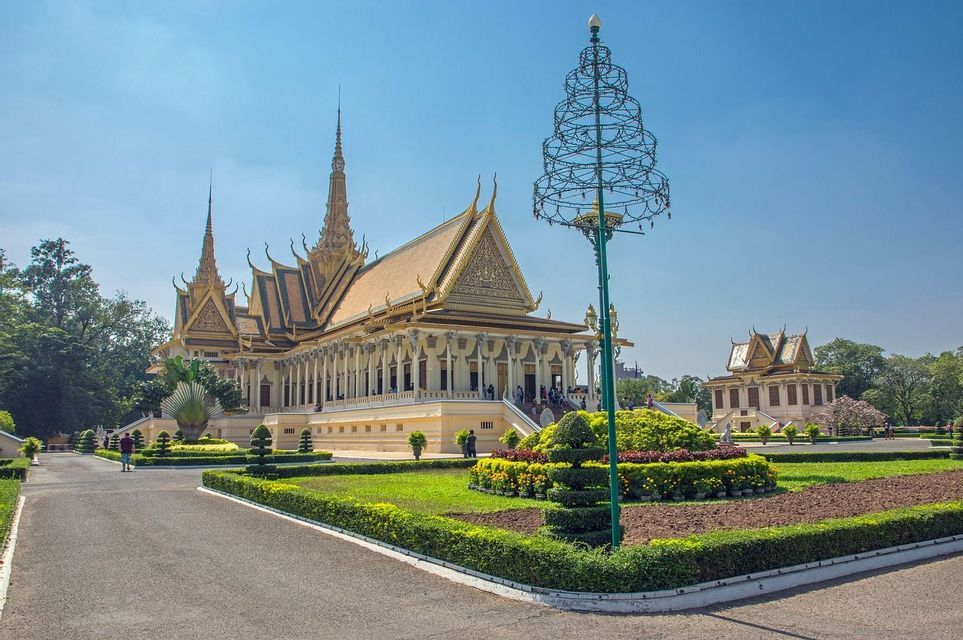 Un palais orné avec un toit doré et des flèches se dresse derrière un jardin bien entretenu doté d'un chemin pavé, sous un ciel bleu clair.