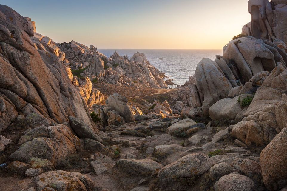 Un sentiero si snoda attraverso un'aspra valle di imponenti formazioni rocciose che portano al mare, illuminato da un tramonto dorato.