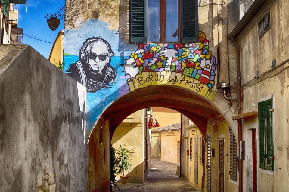 Graffiti colorati, incluse le parole 'Borgo degli Artisti', decorano un arco sopra uno stretto vicolo tra edifici gialli in una giornata di sole.