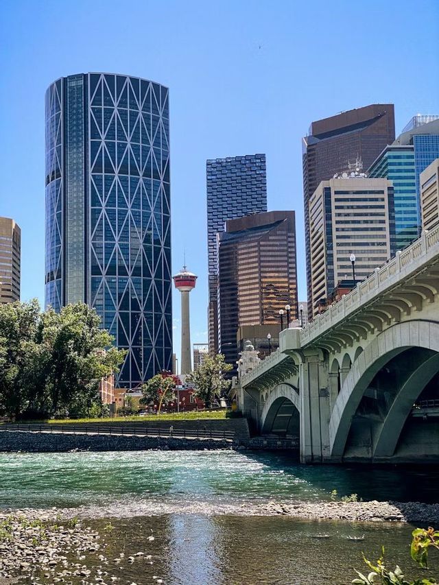 Un horizonte urbano moderno con rascacielos y una torre de observación, visto desde el otro lado de un río con un puente de arco de piedra.