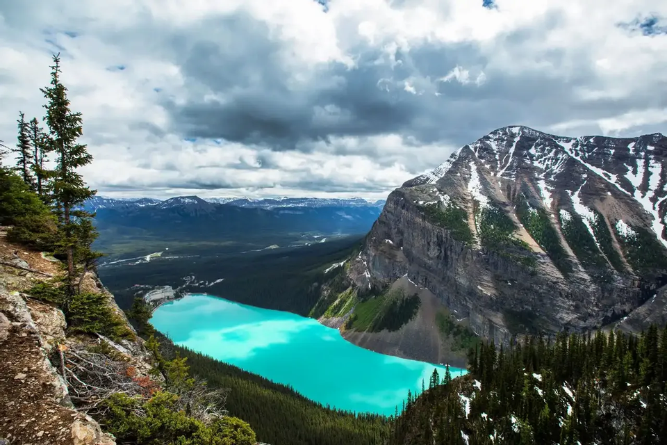 Vista elevada de un lago turquesa brillante, rodeado de bosques de pinos y una gran montaña nevada bajo un cielo nublado.