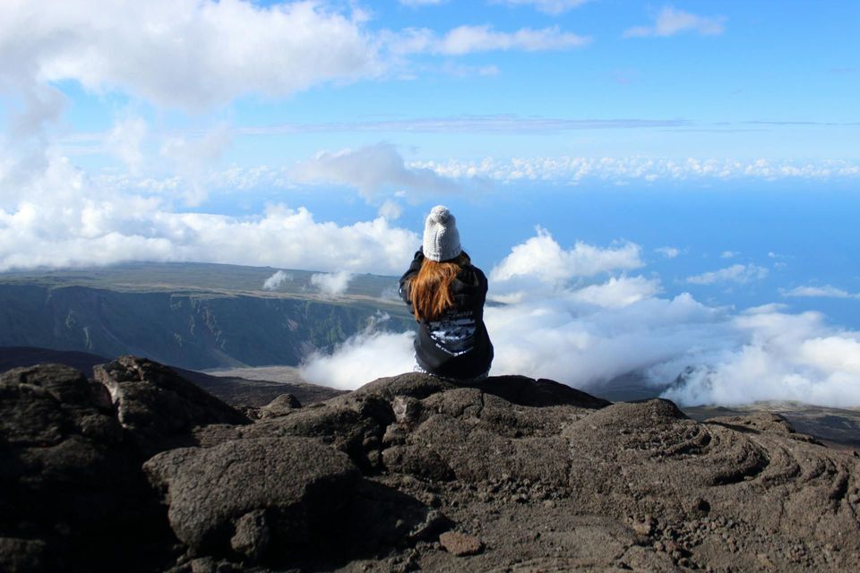 Eine langhaarige Person, von hinten gesehen, sitzt mit einer Mütze auf einem felsigen Gipfel und blickt über eine wolkengefüllte Schlucht.