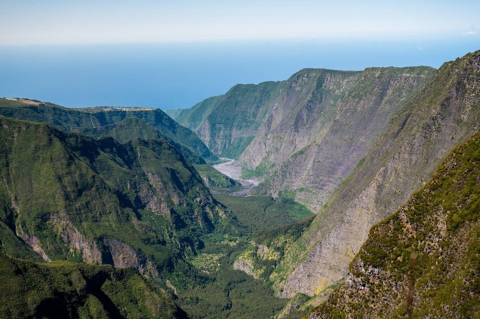 Luftaufnahme eines riesigen, grünen Canyons mit steilen Berghängen, einem Talboden darunter und dem Ozean am Horizont sichtbar.