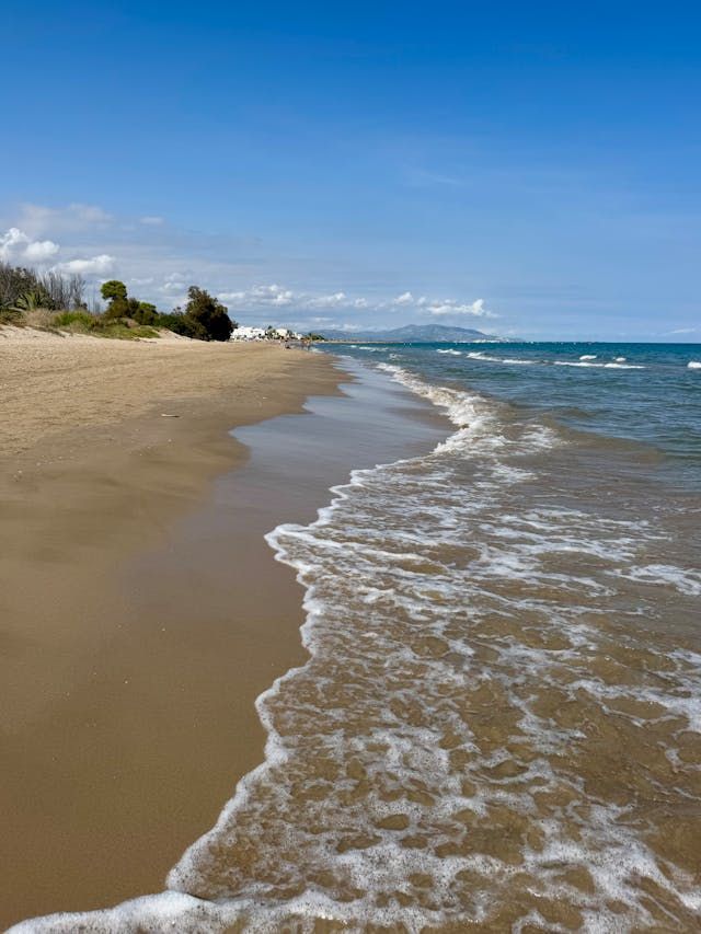 Wellen mit weißem Schaum rollen an einen Sandstrand unter einem klaren blauen Himmel, mit fernen Bergen am Horizont.