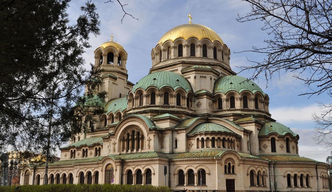 Una cattedrale in pietra riccamente ornata con una grande cupola dorata e diverse cupole verdi più piccole, incorniciata da alberi contro un cielo parzialmente nuvoloso.