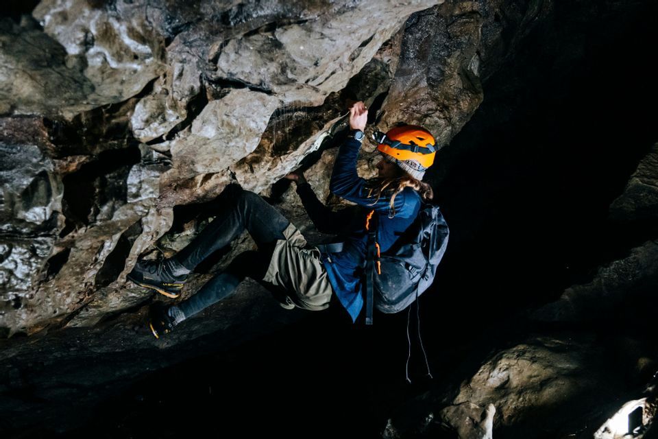 Una persona con un casco naranja y una linterna frontal escala la pared rocosa dentro de una cueva oscura.