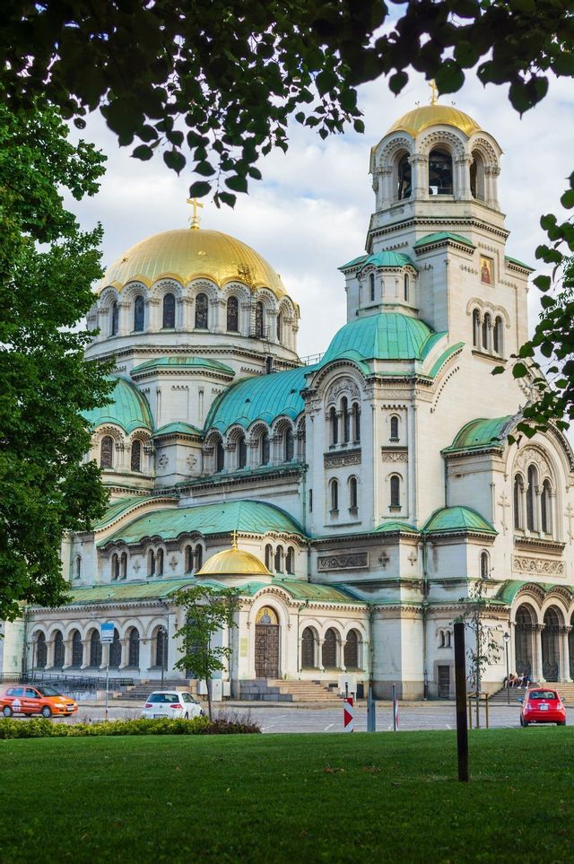 Una grande cattedrale bianca con cupole dorate e turchesi, vista da un prato erboso e incorniciata da foglie verdi.