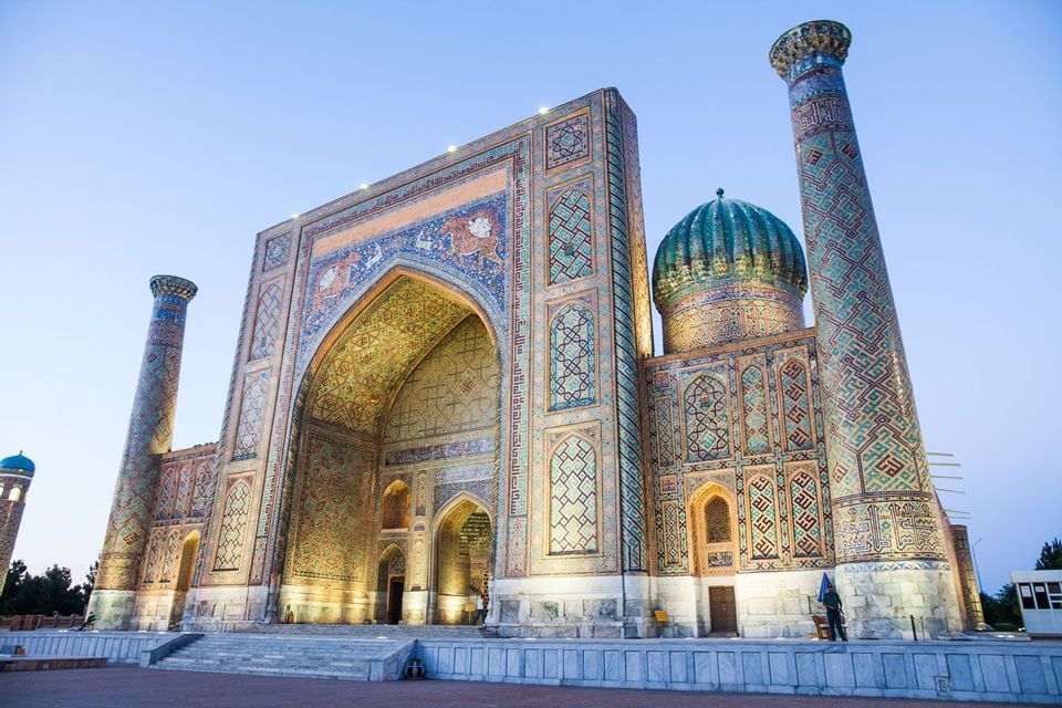 Une médersa ornée de mosaïques complexes bleues et dorées, une grande arche, un dôme turquoise et des minarets, illuminée au crépuscule.