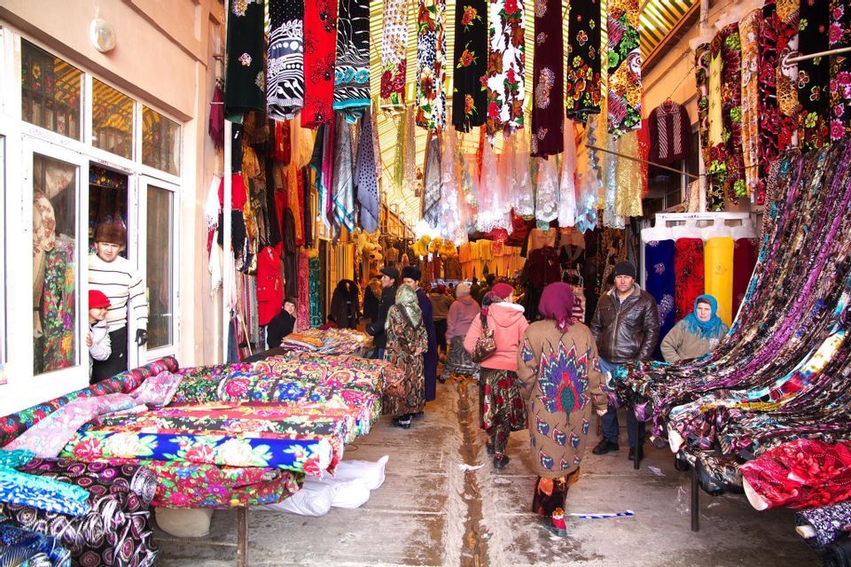A busy outdoor market aisle crowded with people browsing stalls filled with colorful textiles and fabrics.