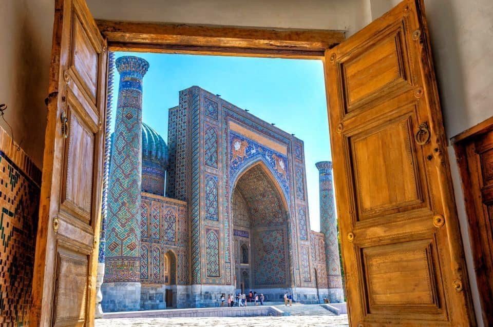 Un edificio ornamentado con intrincados mosaicos azules y dorados y una cúpula, visto desde el interior a través de un conjunto de puertas de madera abiertas.
