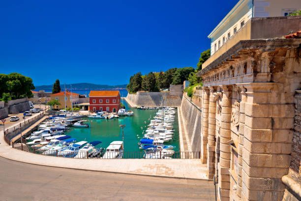A sunny day view of a historic harbor where numerous white boats are docked in turquoise water next to stone fortifications.