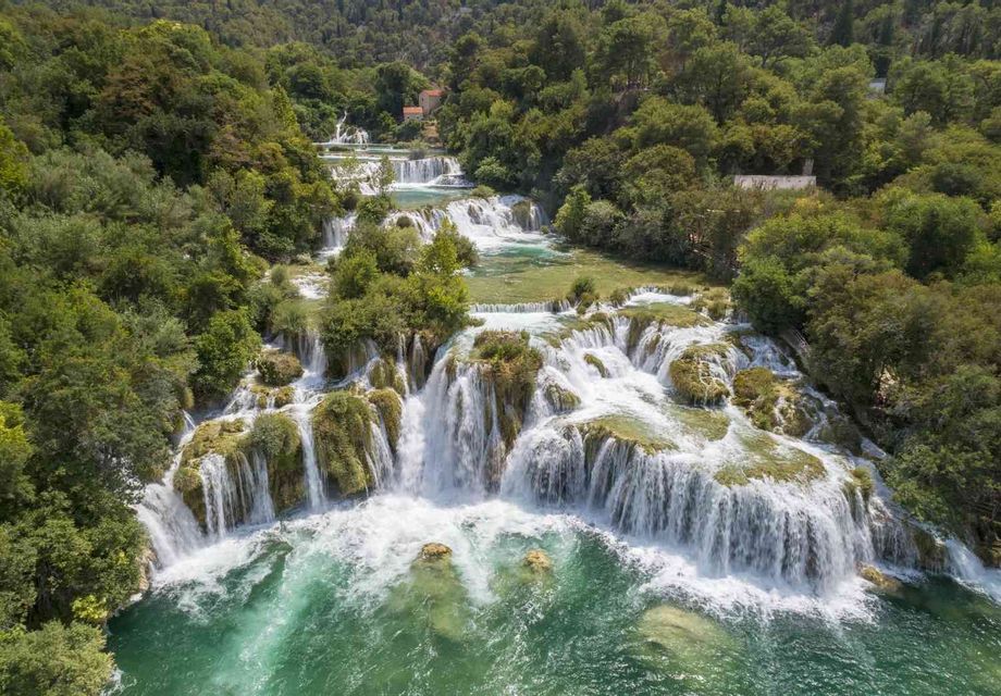 An aerial view of a wide, cascading waterfall flowing into a turquoise river surrounded by a dense green forest.