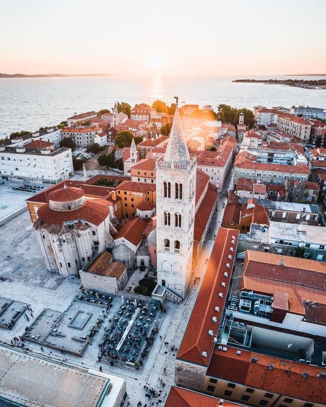 An aerial view of a historic coastal city with a church and bell tower glowing in the golden light of sunset.