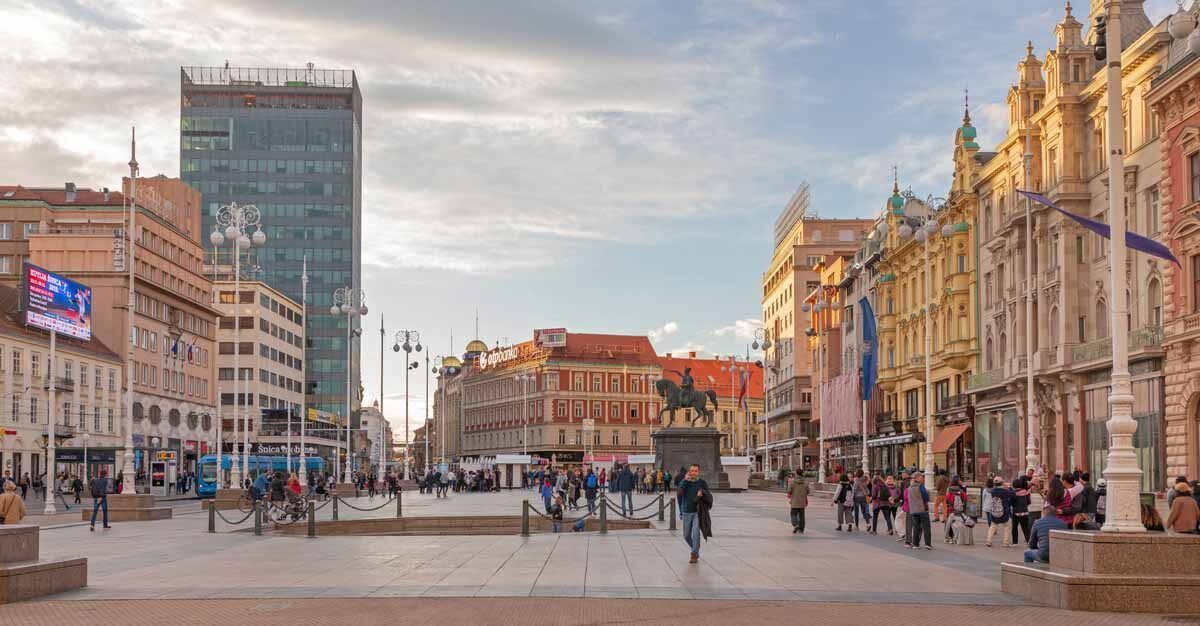 A wide view of a European city square with people walking around an equestrian statue, surrounded by historic and modern buildings under a cloudy sky.