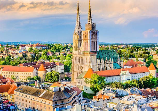 A Gothic cathedral with two spires towers over a cityscape of orange-roofed buildings and lush trees under a partly cloudy sky.
