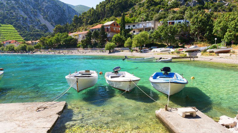 Three small boats are moored in clear turquoise water in a coastal cove, with a village and mountainside in the background.