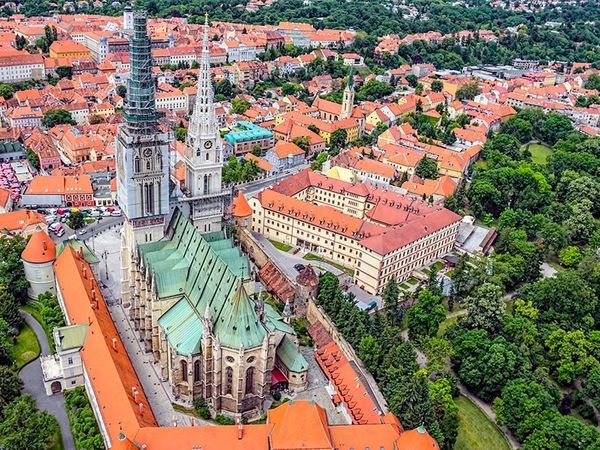 An aerial view of a historic European city, featuring a large cathedral with a green roof and two spires among red-roofed buildings and trees.