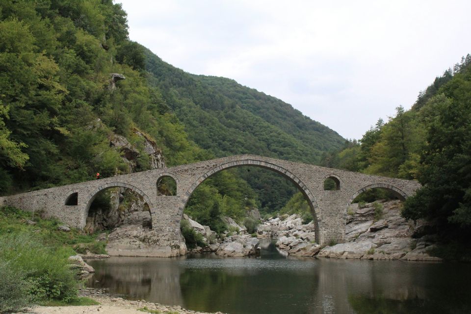 Un pont de pierre à plusieurs arches enjambe une rivière dans une vallée aux collines escarpées et boisées.