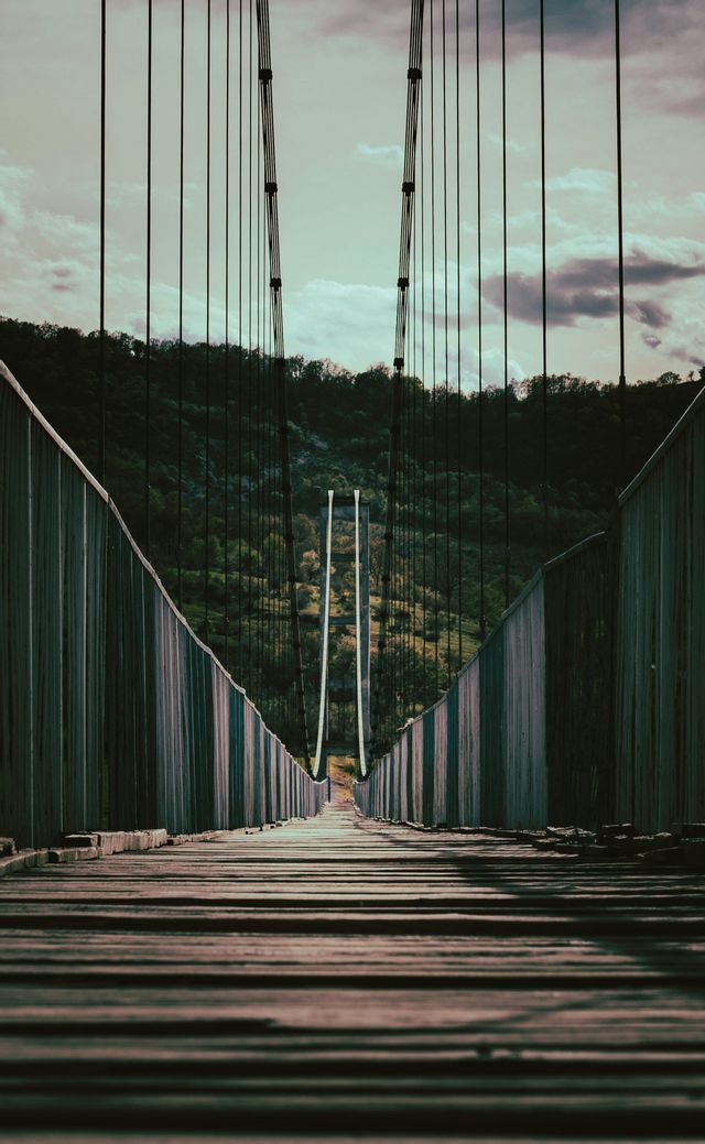 Vue en contre-plongée d'un pont suspendu en bois s'étirant vers une colline verte et boisée sous un ciel nuageux.