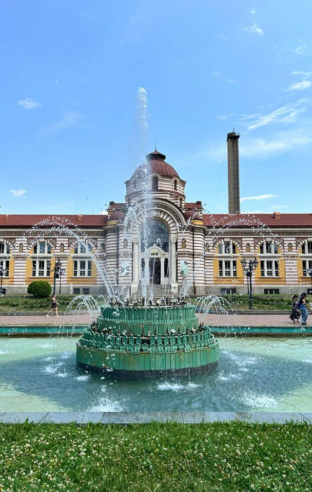 Une fontaine verte à plusieurs niveaux fait jaillir de l'eau sur une place publique, devant un bâtiment orné d'un dôme, sous un ciel bleu clair.