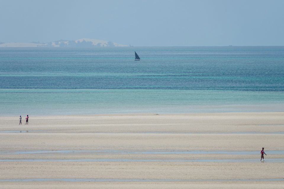 Three people walk on a vast sandy beach at low tide, with a sailboat on the distant blue and turquoise ocean.
