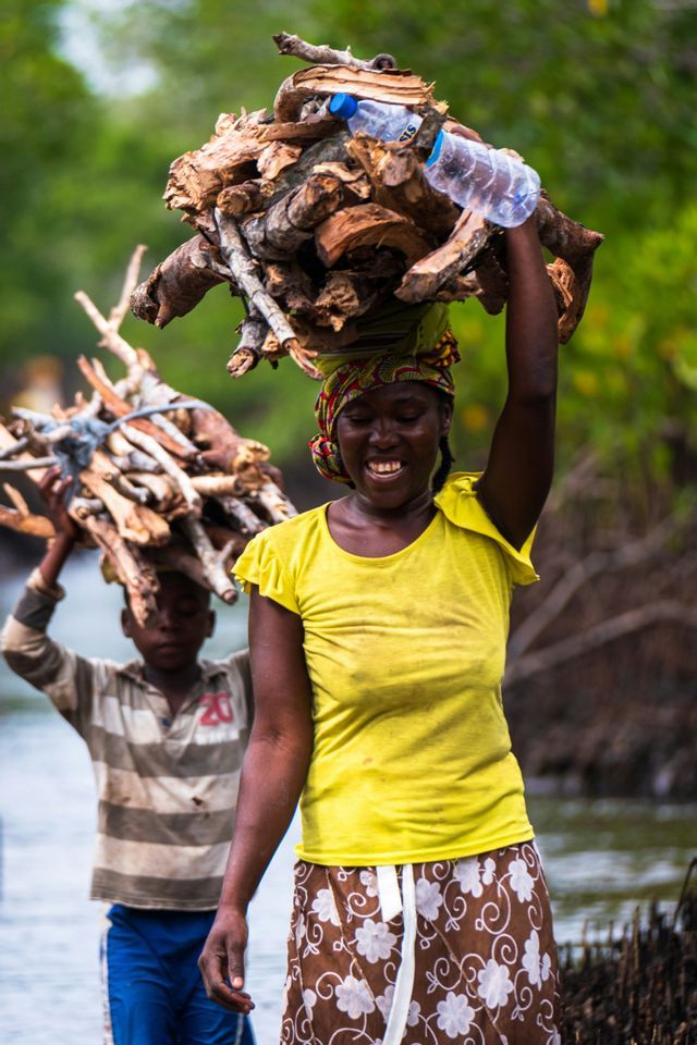A smiling woman in a yellow shirt and a boy walk while carrying bundles of firewood on their heads.