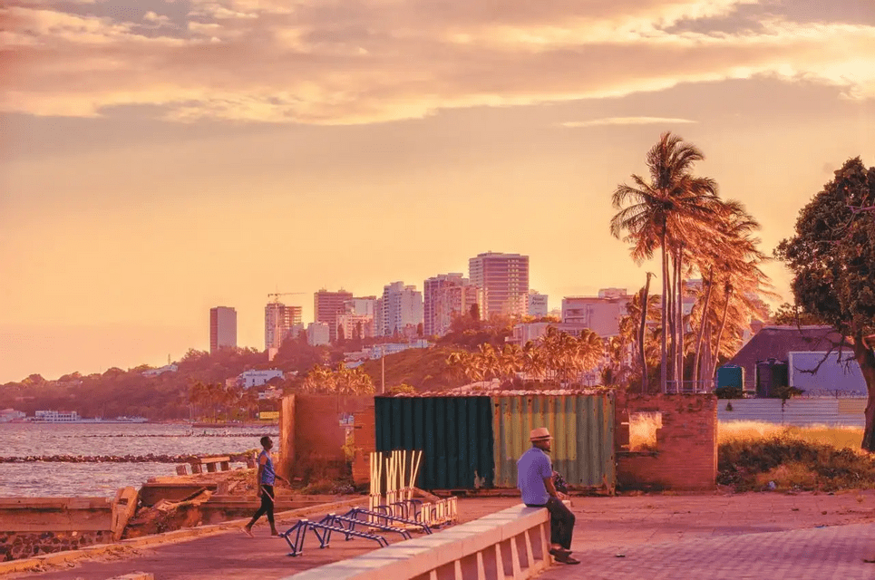 A man sits on a concrete ledge on a waterfront, looking out at a modern city skyline with palm trees during a warm sunset.