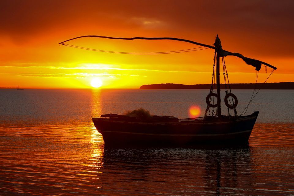 A silhouetted boat floats on calm water during a vibrant orange sunset over the sea.