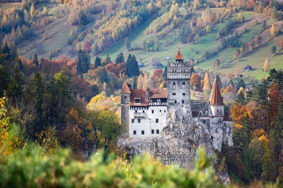 A medieval castle with red-tiled towers stands on a rocky cliff surrounded by hills covered in vibrant autumn trees.