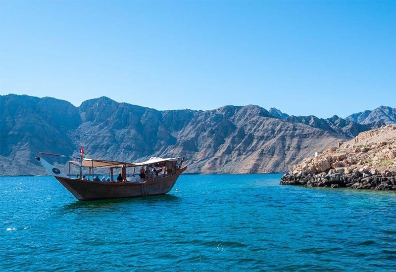 A WeRoad group trip on a traditional Dhow boat floats on turquoise water, with rocky mountains in the background.