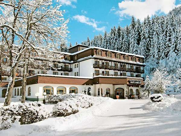 Un edificio alberghiero in stile alpino è circondato da neve alta e da una fitta foresta di pini coperti di neve.