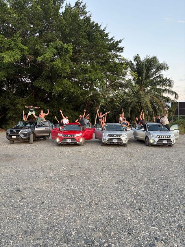Un gruppo WeRoad in viaggio che esulta mentre posa sopra e intorno a quattro auto parcheggiate in un piazzale ghiaioso con alberi rigogliosi sullo sfondo.