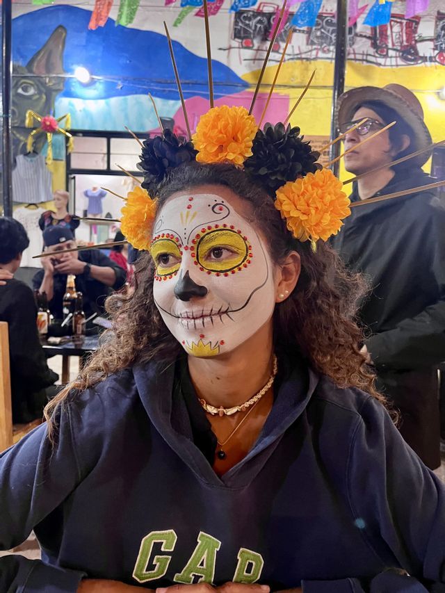 Une femme, le visage maquillé en Calavera, porte une coiffe de fleurs oranges et noires lors d'une célébration en intérieur.