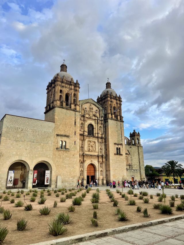 Une église en pierre ornée, dotée de deux clochers, vue depuis une place avec de petites agaves, sous un ciel nuageux.