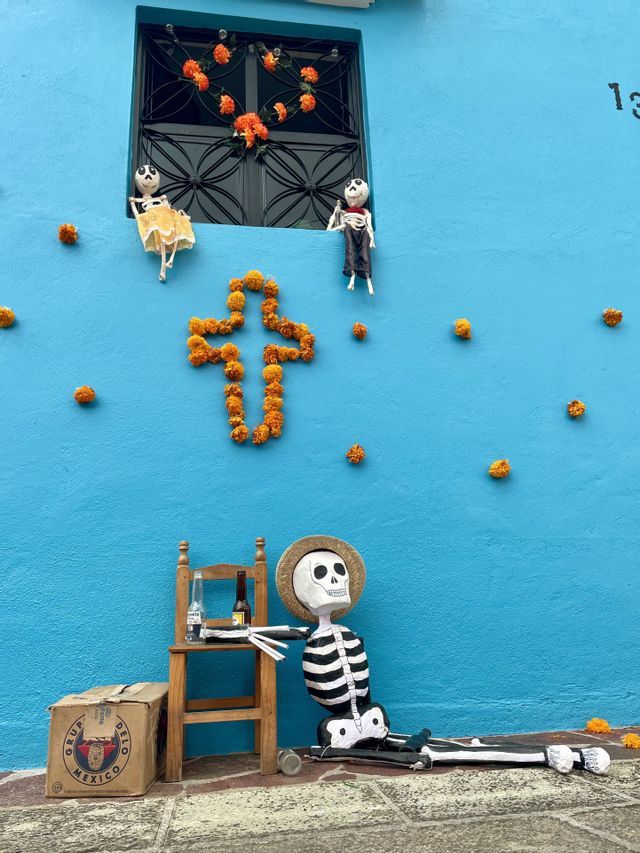 Une grande figurine de squelette est assise par terre contre un mur bleu, orné d'une croix de fleurs de souci et de petites figurines de squelettes.